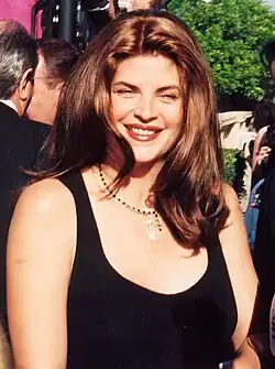 Kristie Alley (1994) lächelt mit langen braunen Haaren, die ein schwarzes ärmelloses Oberteil trägt. Sie hat eine Halskette mit einem Anhänger. Im Hintergrund sind mehrere Personen teilweise sichtbar, sowie grüne Landschaftselemente. Das Foto wurde bei den Emmys 1994 im Freien aufgenommen.