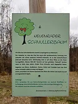 Text auf der Tafel: „Neuenrader Schnullerbaum. Die Idee des Schnullerbaumes stammt aus Dänemark. Der Gedanke ist, dass das Kind die sonst eher problematische Trennung vom Schnuller mit einem positiven Erlebnis verbindet, da es den Schnullerbaum jederzeit besuchen kann. Gleichzeitig wird es auf diese Weise auch an die Natur herangeführt. Ebenso fällt der Verzicht auf den geliebten “Schnulli” leichter wenn es sieht, dass auch andere Kinder ihren Schnuller abgegeben haben. (Sic! Kommafehler nach „leichter“ im Original) Begleitet von Eltern, Großeltern, Tanten, Onkel und Freunden kann das Kind seinen Schnuller selbst an einen Ast hängen. Meist unterstützt ein kleines Geschenk der Eltern die Aktion und macht sie zu einem unvergessenen Ereignis. (Vom vorhergehenden Text abgesetzt und in anderer Farbe:) Achtung! Bitte verwenden Sie aus Naturschutzgründen nur neutrale Bindfäden zur Aufhängung. Keine Schleifenbänder, Kartons oder sonstiges Dekomaterial. Wir müssten sonst den Schnuller entfernen. forumneuenrade e. V. Abteilung Stadtverschönerung“