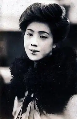 Die schwarz-weiß Fotografie ist im Hochformat und zeigt Urabe Kumeko in die Kamera blickend. Sie hat dunkles Haar das schwungvoll zusammen gesteckt ist. Sie trägt einen dunklen flauschigen Schal.