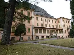 Das stillgelegte Kurhotel von Acquarossa ist von hohen Bäumen umgeben. Das imposante Gebäude hat zwei Seitenflügel und einen nach hinten gerückten Gebäudekorpus in der Mitte. Das Gebäude ist leicht rosa und die Fensterläden sind olivgrün. Alle Läden sind geschlossen. Der Garten und das Gebäude sind nicht heruntergekommen, aber ungepflegt.