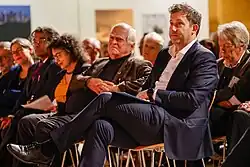 Eine Gruppe von Menschen, die bei einer Veranstaltung in einem Gebäude sitzen und sich aufmerksam konzentrieren. Ein Mann in einem Anzug im Vordergrund sitzt mit überkreuzten Beinen und vermittelt einen engagierten und formellen Eindruck.