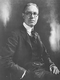 Älterer Mann von leicht seitlich vorn, sitzend und den Kopf zur Kammer gewandt, im dunklen Anzug, dunkler Weste, dunkler Krawatte und weißem Hemd und die linke Hand in der Hosentasche in schwarzweiß.