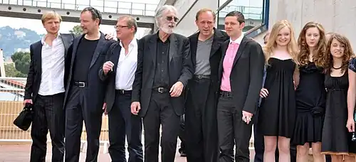 Das Bild zeigt die Darsteller und den Regisseur des Films Das weiße Band bei den Filmfestspielen in Cannes 2009. Die Gruppe besteht aus neun Personen, die elegant gekleidet sind. Die Männer tragen klassische Anzüge, während die Frauen in stilvollen schwarzen Kleidern erscheinen. Sie stehen zusammen vor einem modernen Gebäude mit großen Glasfenstern und einem Geländer im Hintergrund. Die Atmosphäre wirkt festlich und professionell, passend zu einem renommierten Filmfestival. Von links: Michael Kranz, mit blonden Haaren in weißem Hemd und schwarzer Hose, Rainer Bock mit dunklem T-Shirt und Jacke, Burghart Klaußner mit Brille und grauem Sakko, Michael Haneke mit weißem Bart und Sonnenbrille in schwarzem Anzug, Ulrich Tukur mit Glatze und grauem T-Shirt, Christian Friedel mit dunklem Anzug und rosa Hemd, Maria-Victoria Dragus mit langen blonden Haaren in schwarzem Kleid, Roxane Duran mit langen braunen Haaren in schwarzem Kleid, Janina Fautz mit lockigen braunen Haaren in schwarzem Kleid. Alle schauen zur Kamera und lächeln leicht. Sie posieren für ein Gruppenfoto, einige mit entspannten Gesichtsausdrücken, andere mit einem leichten Lächeln.