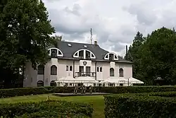 Das Waldhotel Peršlák, ein dreistöckiges Gebäude mit rosa-weißer Fassade eingebettet zwischen Bäumen. Auf dem Gebäude ist ein Wappen mit Doppeladler, im kleinen Park vor dem Gebäude stehen zwei Kanonen.