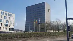 Moderne städtische Szene mit zwei markanten Gebäuden, die zusammen ein Büro- oder Verwaltungszentrum darstellen: Rechtes Gebäude: Es ist ein hohes, rechteckiges Bauwerk mit einer grauen Fassade, die in klaren Linien gestaltet ist. Die Fenster sind in mehreren horizontalen Reihen angeordnet. Oben rechts an der Fassade befindet sich ein auffälliges, goldfarbenes Logo in der Form eines stilisierten „G“. Linkes Gebäude: Dieses Bauwerk ist kleiner und hat eine weiße Fassade mit großen Fenstern, die in einem gleichmäßigen Raster angeordnet sind. Es wirkt offen und modern, im Gegensatz zum eher massiven Stil des rechten Gebäudes und hat einen gelben Balkon. Aglaia Konrad entwarf den weithin sichtbaren gelben Balkon am Provinzhaus der Provinz Flämisch-Brabant. Umgebung: Im Vordergrund sind fünf Fahnenmasten mit verschiedenen Flaggen zu sehen. Darunter befindet sich die belgische Nationalflagge sowie andere regionale oder organisatorische Flaggen. Die Umgebung ist von einem klaren Himmel mit blauem Hintergrund geprägt, und im unteren Bereich des Bildes sind einige kahle Bäume und ein gepflegter Grünstreifen sichtbar