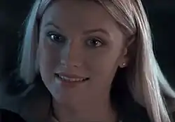 Lily Travers mit langen, hellen Haaren, die sanft über die Schultern fallen. Die Haare stehen im Kontrast zum dunklen Hintergrund, wodurch sie besonders hervorgehoben werden. Lily Travers lächelt freundlich in die Kamera. Sie trägt ein dunkel gefärbtes Kleidungsstück, das jedoch keine spezifischen Details aufweist, da das Licht den Fokus auf die Haare und den Kopf legt. Der dunkle Hintergrund ist neutral und gleichmäßig, ohne erkennbare Muster oder Objekte.