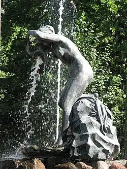 Foto einer Statue einer nackten Frau, die leicht vornübergebeugt steht und einen Krug ausgießt. Aus dem Krug fließt echtes Wasser. Hinter ihr ist vor grünen Büschen und Bäumen der Wasserstrahl einer Fontäne zu sehen.