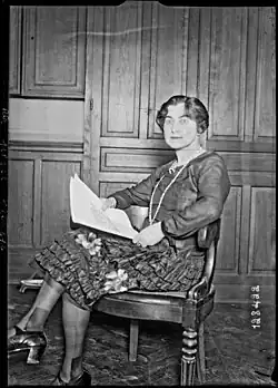 Die Schwarz-Weiß-Fotografie zeigt eine auf einem Holzstuhl sitzende Frau in einem Kleid; sie hat die Beine übereinandergeschlagen, hält einen Papierbogen in den Händen und blickt mit wachen Augen in die Kamera.