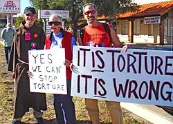 Louis Vitale, Megan Rice und Jim Haber bei einem Protest bei Ft. Huachuca, Arizona. Der Pater trägt das Mönchshabit und die drei tragen zusammen zwei große Plakate mit Protest-Slogans.