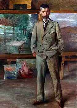 Ein Gemälde mit Eichfeld in seinem Atelier im Vollporträt. Er steht vor einigen seiner Bilder und trägt einen grauen Anzug. Das Haar ist dunkel und an den Schläfen grau, er trägt zudem einen gestutzten Vollbart.