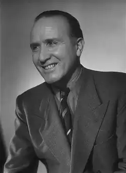 Lächelnder Luccioni Harcourt (1947) auf dem schwarz-weiß Portrait, trägt einem gut sitzenden Anzug mit Krawatte. Der Anzug hat ein klassisches Revers mit einer Tasche auf der linken Seite. Die Krawatte ist gestreift. Der Hintergrund ist schlicht und neutral, sodass der Fokus auf der abgebildeten Person liegt.