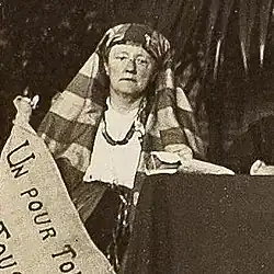 Lucy Thoumaian ist an einem Tisch sitzend zu sehen. Sie hält ein Plakat das vom Bild abgeschnitten wird. Sie trägt eine Kopfbedeckung und traditionelle armenische Kleidung.