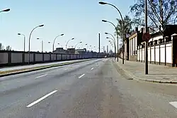 Seitliche Farbfotografie von einer Straße mit einer langen Mauer auf der linken Seite und einer Fabrikfassade auf der rechten Seite. Im Hintergrund fährt ein Auto und hinter der linken Mauer sind Häuser und ein Fabrikschornstein.