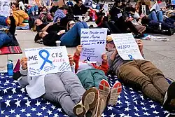 Mehrere Personen demonstrieren im Rahmen einer „LiegendDemo“ am 11. Mai 2023 in Berlin. Im Vordergrund liegen drei Personen mit Schildern in den Händen. Auf dem ersten Schild steht: „Wir wollen unser Leben zurück! ME/CFS nimmt alles. Ruben, 14 J., moderat seit 03/22. Lena, 40 J., schwer betroffen seit 07/22. Cordula, 44 J., moderat betroffen, erkrankt 12/20.“ Auf dem Schild der Person in der Mitte steht: „Mein Bruder und meine Mama sollen endlich wieder gesund sein. Wir brauchen Forschung, Versorgung, Anerkennung.“ Auf dem dritten Schild steht: „Jetlag, Grippe, Hangover gleichzeitig. Und jetzt lauf mal einen Marathon! So in etwa fühlt sich ME/CFS an. Ein täglich niemals endender Alptraum für dich und deine Familie.“ Im Hintergrund sieht man weitere Personen, die im Liegen demonstrieren.