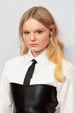 Maria Dragus mit langen, welligen blonden Haaren, trägt ein weißes Hemd mit schwarzer Krawatte und ein schwarzes Korsett darüber, schaut ernst in die Kamera, vor hellem Hintergrund auf der Medienboard Party bei der Berlinale 2019.