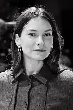 Hannah Gross bei der Vorstellung des Films The Adults auf der Berlinale 2023 in einer Schwarz-Weiß-Aufnahme, die von den Schultern aufwärts fotografiert wurde. Sie trägt ein Hemd mit Kragen und Knöpfen sowie Ohrringe. Ihre Haare sind schulterlang. Der Hintergrund ist unscharf, was darauf hindeutet, dass das Foto in einem geschlossenen Raum aufgenommen wurde.