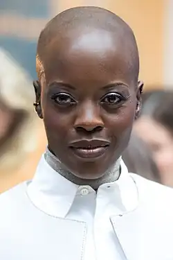 Florence Kasumba mit glattem Kopf und elegantem weißen Oberteil mit hohem Kragen und auffälligen Knopfdetails, lächelt in die Kamera beim Empfang des FilmFernsehFonds Bayern zur Berlinale 2024. Der Hintergrund ist leicht verschwommen, deutet aber auf eine Innenraumkulisse mit weiteren anwesenden Personen hin.