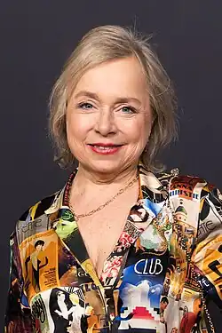 Christine Urspruch mit blondem Haar. Sie trägt ein auffälliges Hemd mit buntem Muster, das verschiedene Bilder und Textelemente enthält. Zu den erkennbaren Texten auf dem Hemd gehören Wörter wie „CLUB,“ „GOU,“ „ANNA,“ „PLACE,“ „GERTIE,“ und „CHRISTIE.“ Christine Urspruch trägt außerdem eine goldene Halskette. Der Hintergrund des Bildes ist dunkel gehalten und entstand beim NRW-Empfang der Berlinale 2024.