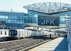 Panorama-Blick von einem Bahnsteig auf eine brückenartiger Überbauung über mehreren Gleisen mit einem Zug der Linie A. Überbauung ist beschriftet mit übergroßem Schriftzug „Howard Beach JFK“, hat eine Fassade aus Glas und Aluminium und eine Dachkonstruktion teilweise aus Raumfachwerk.