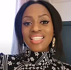 Maïmouna Doucouré sitzt vor einer beige-schwarzen Wand. Sie trägt ein schwarzes Oberteil mit schwarz-weißen Streifen und weißen Blütenaufdrücken, an jedem Ohr je einen Ohrstecker, an dem drei flache, gelbbraune Steine hängen, sowie ihre schwarzen Haare als strähnige, bis zum Hals reichende Bobfrisur.