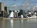 Mittig im Bild befindet sich das Science Center umgeben von einer Skyline aus Hochhäusern. Vordergründig ist Wasser zu sehen.