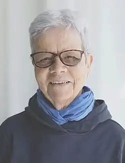 Madeleine Marti schaut leicht lächelnd in die Kamera. Sie hat kurzes helles Haar, trägt eine schwarze Kunststoffbrille und hat ein hellblaues Tuch um den Hals. Ihr Pullover ist dunkelblau und der Hintergrund hell.