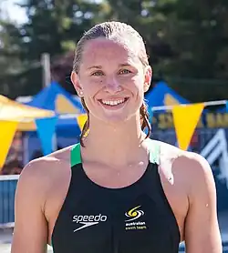 Madeline Groves befindet sich neben dem Pool unter freiem Himmel. Sie hat nasse Haare und trägt einen schwarzen Badeanzug mit grünem Schulterträger. Der Badeanzug zeigt die Aufschrift des Ausrüsters und die Worte australian swim team in Kleinschreibung. Im Hintergrund sind gelbe und blaue Wimpel zur Markierung der Tauchzone erkennbar.