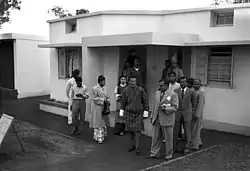 Kesang Choden in einer Schwarz-Weiß-Aufnahme von 1954. Sie steht hinter ihrem Mann, Jigme Dorji Wangchuck, der zusammen mit 12 anderen Würdenträgern aus einem Flachdach-Gebäude tritt.