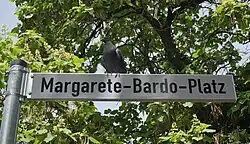 Weißes Straßenschild das den Namen Margarete-Bardo-Platz trägt. Auf dem Schild sitzt mittig eine graue Taube und im Hintergrund ist ein Zigarrenbaum zu sehen.
