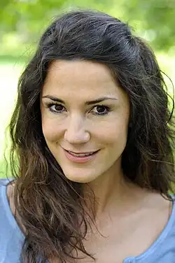 Mariella Ahrens 2013 Portrait vor grünen Hintergrund ausgeblendet, blauem u-förmig ausgeschnittenen Shirt mit schwarzen langem Haar links nach vorne fallend, rechts nach hinten gekämmt