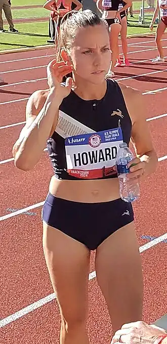 Läuferin ab Kniehöhe im Laufdress auf einer Aschenbahn mit Wasserflasche in der Hand von vorn.