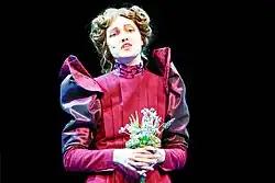 Marta Kizyma in einer historischen Kostümierung auf der Bühne des Maxim Gorki Theaters in der Inszenierung von Der Untertan (2023). Sie trägt ein tiefrotes Kleid mit aufwendig gestalteten, gepufften Ärmeln und detaillierten Verzierungen am Kragen. In ihren Händen hält sie einen kleinen Strauß aus zarten Blumen. Der Hintergrund ist dunkel gehalten.