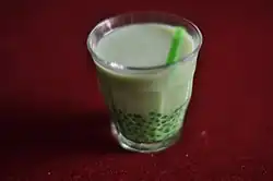 Ein transparentes Glas mit grün bedruckten Blättern beinhaltet eine hellgrüne Flüssigkeit. Es steckt ein dunkelgrüner Strohhalm im Glas, das auf dunkelrotem Untergrund steht, der das gesamte Bild ausfüllt.