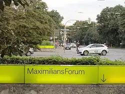 Straßenszene, im Vordergrund eine Reihe gelber Leuchtbausteine mit schwarzer Aufschrift „MaximilansForum“ und Pfeil nach unten