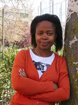 Melibea Obono. Afrikanerin mit ungezähmten Haaren und einem orangefarbenen Oberteil lehnt an einem Baum.