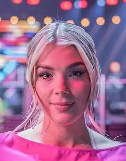 Keyyo beim Melodifestivalen 2025, hat mittellanges blondes Haar, das in der Mitte gescheitelt ist und leicht über die Schultern fällt. Sie trägt ein rosafarbenes Oberteil. Der Hintergrund ist verschwommen, er zeigt bunte Lichtstreifen und punktuelle Lichter, die auf eine Bühne oder Veranstaltung hinweisen könnten. Die Beleuchtung des Bildes ist überwiegend in rosa und blauen Tönen gehalten, was eine lebendige, aber verträumte Atmosphäre schafft.