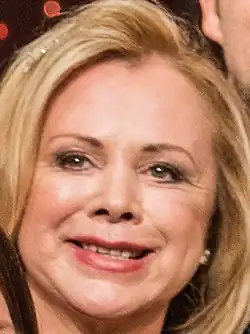 Marijke Amado (2018) Portraitausschnitt aus einem Gruppenbild der Geburtstagssendung zum 70ten von Jürgen von der Lippe. Sie lächelt mit blonden mit Volumen gestylten Haaren und eleganter Kleidung, posiert sie in der Gruppe. Der Hintergrund ist dunkel und zeigt unscharfe Lichter, die auf die Studiokullisse hindeuten könnten.