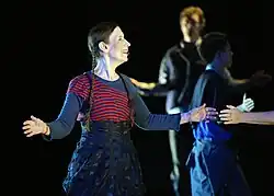 Ein Bühnenausschnitt zeigt Meredith Monk in einem Kleid und ausgebreiteten Armen singend.