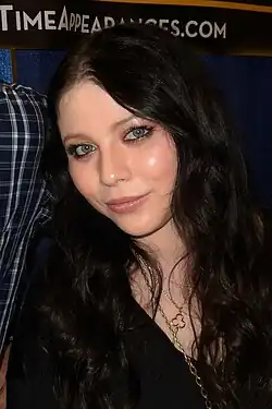 Michelle Trachtenberg Foto Portrait, schwarze lange Haare mit einer goldenfarbenen Kurz-Kette und einer Langkette mit größeren Kettengliedern, gekleidet in schwarz, einer weiteren Person nur Ansätze vom Arm sichtbar in einem karierten Hemd ist unkenntlich ausgeschnitten im Hintergrund ist ein Banner mit einem Schriftzug time appearances.com erkennbar