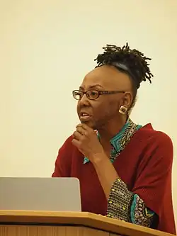 Michelle Parkerson sitzt an einem Rednerpult. Sie trägt einen dunkelroten Pullover mit bunten Ärmeln und Kragen, eine dunkelbraune, ovale Brille sowie einen quadratischen, weißen Ohrstecker mit einer runden, goldenen Mitte am linken Ohr. Ihre schwarzen Haare trägt sie an den Seiten rasiert und am Oberkopf als kurze Locken.