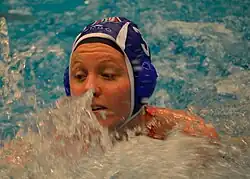 Mieke Cabout während eines Spiels. Zu sehen ist außer spritzendem Wasser die linke Schulter, das Gesicht von Cabout und die blaue Badekappe mit der Nummer 3.