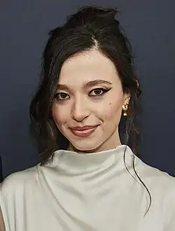 Mikey Madison posiert lächelnd bei den 2024 Toronto International Film Festival, die ein hochgeschlossenes, weißes Kleidungsstück trägt. Ihr dunkles Haar ist elegant in einer Hochsteckfrisur gestylt, wobei einige lockere Strähnen das Gesicht umrahmen. Sie trägt goldene Ohrringe, die subtilen Glanz verleihen. Der Hintergrund ist einheitlich dunkel.