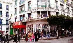 Eine Straßenaufnahme die die Milk Bar bei Tageslicht und mit vielen Menschen, die davor stehen, zeigt.