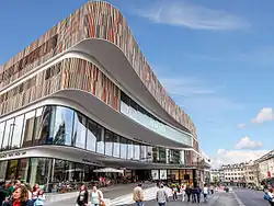 Das Bild zeigt ein modernes Gebäude, das Minto in Mönchengladbach, mit einer auffälligen, geschwungenen Fassade, die aus vertikalen Holzlamellen besteht. Diese Lamellen verleihen dem Gebäude eine elegante und zeitgenössische Optik. Große Glasfenster ziehen sich über mehrere Stockwerke, wodurch das Innere sichtbar wird und das Gebäude ein offenes und einladendes Erscheinungsbild erhält. Im Erdgeschoss befinden sich Geschäfte und Restaurants, unter anderem ein Restaurant der Kette „Vapiano“. Vor dem Restaurant sind Tische und Stühle im Freien aufgestellt, wo Menschen gemütlich sitzen. Weitere Personen gehen auf der belebten Straße vor dem Gebäude. Der Himmel ist blau, durchzogen von ein paar Wolken.