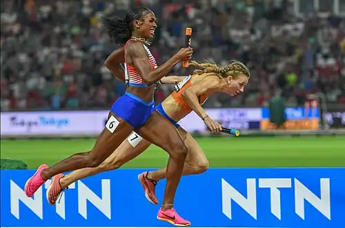 Alexis Holmes in rot-weiß mit Staffelstab in der Hand zieht an Femke Bol in Orange, die stolpert und die Kontrolle über den Staffelstab verliert, vorbei zu WM-Gold.