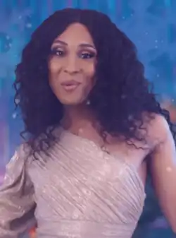 Michaela Jaé Rodriguez steht vor einer dunkelblauen Wand mit aufgemalten Schneeflocken und einer Art Polarlicht. Sie trägt ein silbernes Kleid, das ihren linken Arm freilässt, und ihre schwarzen Haare als Locken.