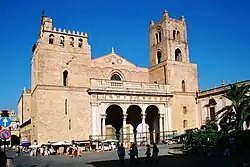 Frontale Farbfotografie eines Gebäudes mit zwei breiten Ecktürmen und einem Giebeldach in der Mitte. Über den Eingangsbögen ist ein Balkon mit weißem Steingeländer und an der Fassade sind symmetrisch Bögen eingemeißelt. Am linken Turm hängen in vier Rundbögen Glocken und an der Fassade sind wenige Fensteröffnungen zu sehen. Um das Gebäude stehen Verkaufsstände.