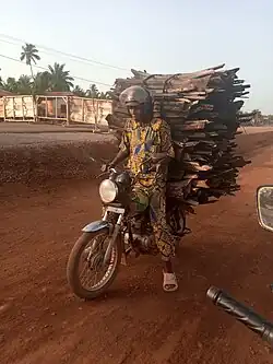 Am Rande von Katagon in Benin, nahe der Fernstraße RNIE1, stoppt ein Brennholz-Transporteur sein Motorrad, der Brennholzstapel aus gut ein Meter langen, dicken Ästen auf dem Sozius überragt den Fahrer im Stand (2020)