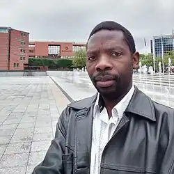 Selfie eines mittelalten, neutral blickenden Mannes auf einem Stadtplatz. Er trägt ein weißes Hemd und eine schwarze Lederjacke.