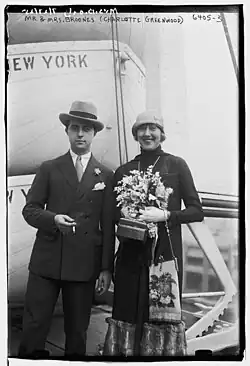 Mr. & Mrs. Broones (Charlote Greenwood) Schwarz-Weiß-Fotografie von dem Ehepaar Broones, die nebeneinander stehen. Martin Broones auf der linken Seite trägt einen Anzug mit Krawatte und Hut. In ihrer rechten Hand hält er eine Zigarette. Charlote Greenwood auf der rechten Seite hat ein langes Kleid an, dazu einen Schal und ebenfalls einen Hut. Sie hält in der linken Hand einen Blumenstrauß und in der rechten eine kleine Handtasche. Im Hintergrund des Bildes befindet sich ein Rettungsboot mit der Aufschrift „NEW YORK“. Oberhalb des Bildes ist handschriftlich der Text „MR. & MRS. BROONES (CHARLOTTE GREENWOOD) 6405-3“ vermerkt.